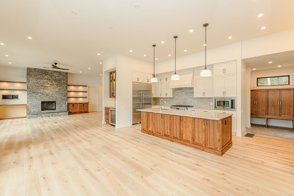 Modern open-concept kitchen and living space in a custom Cowichan Valley home, featuring natural wood cabinetry, wide-plank hardwood floors, and a stone fireplace—showcasing craftsmanship and luxury home design on Vancouver Island.