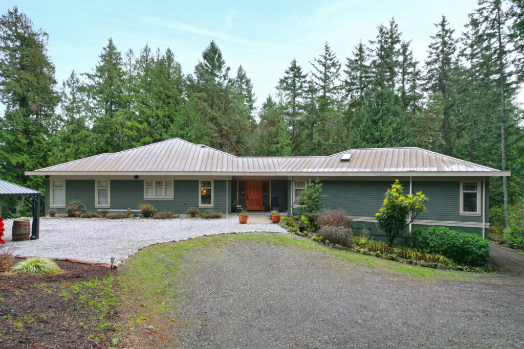 Front view of a single-story custom home surrounded by forest in the Cowichan Valley, featuring a modern metal roof, wide horizontal layout, and natural landscaping